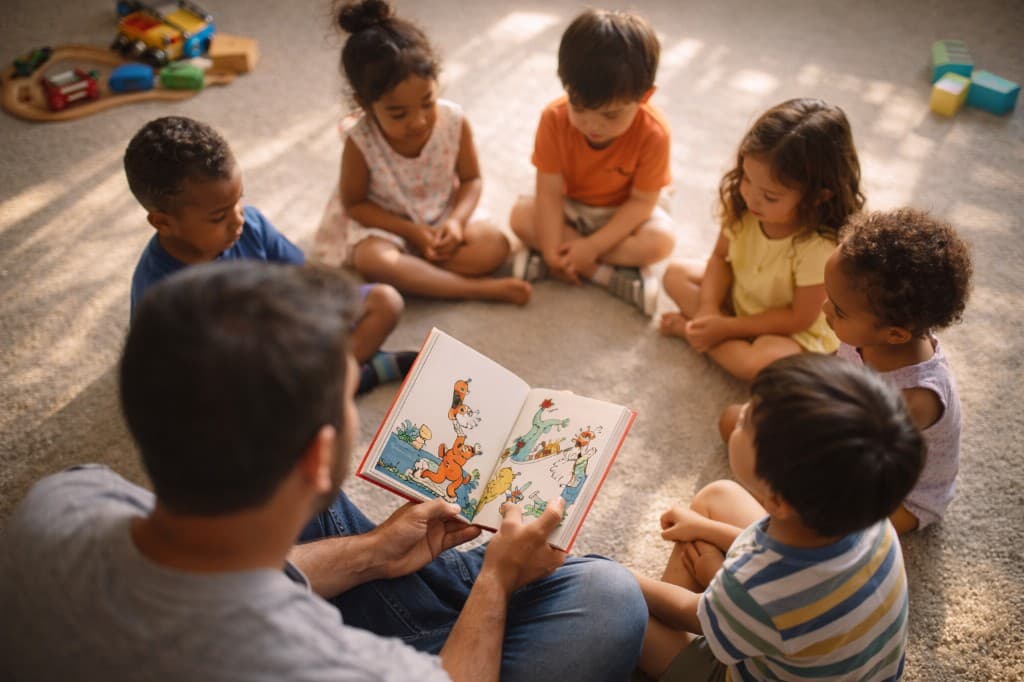 A teacher reads a colorful picture book to a diverse group of young children sitting in a circle on a carpeted floor in a sunlit room.